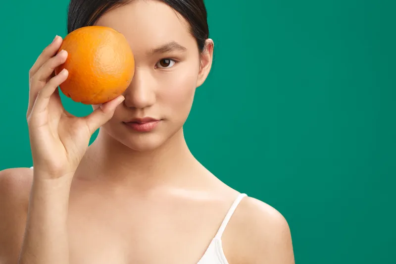 asian woman holds orange near eye in studio setting with a green background, soft light, and composed gaze, creating a fresh, calm and modern portrait.