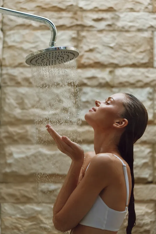 young woman enjoying warm outdoor shower with stone wall in background, wet hair, relaxed mood, natural light, wellness concept, summer day