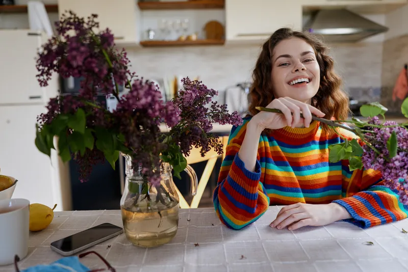 Smiling woman in a multicolor sweater sits at a kitchen table with flowers and fruit, bright natural light, cozy home setting, candid moment of everyday cooking and relaxation, warm mood.