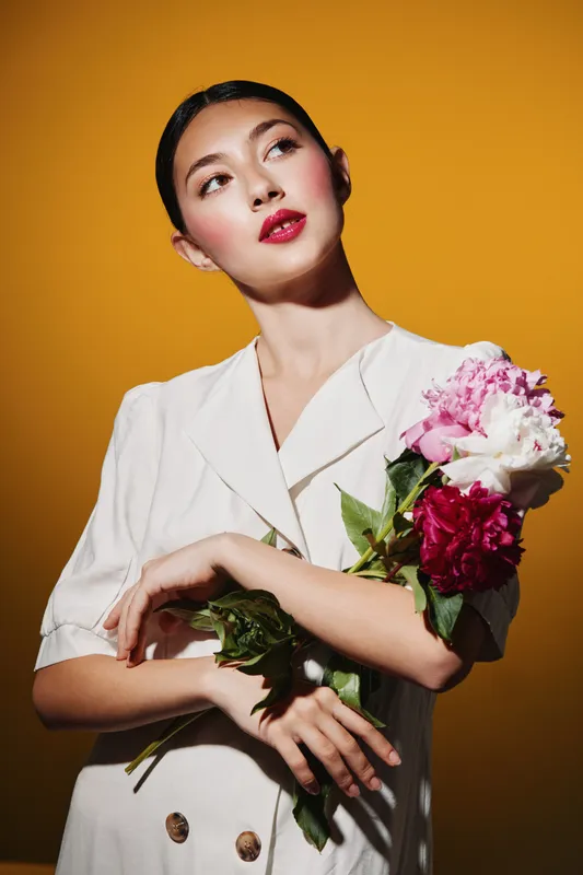 Elegant young woman with red lipstick holding a colorful bouquet, wearing a white blazer against orange background. Creative beauty and fashion concept with sophisticated style.