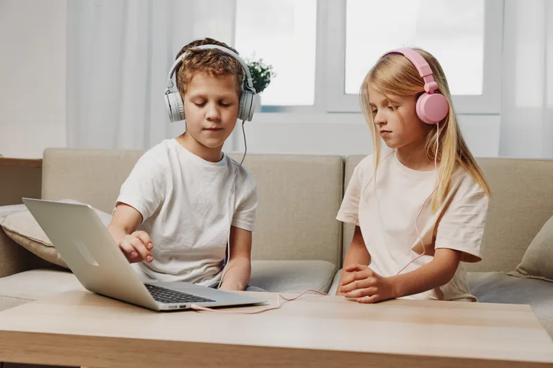 Two children wearing headphones sit on a couch using a laptop, focused on screen. Modern home interior with natural light creates a calm learning and entertainment environment.