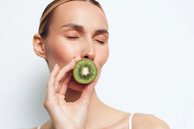 Closeup of woman with closed eyes holding fresh green kiwi slice near lips, soft natural light, smooth skin, minimal makeup, white background, healthy lifestyle concept