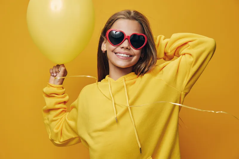 Smiling woman wearing red heart-shaped sunglasses and yellow hoodie holding yellow balloon with cheerful expression on yellow background. Celebration, happiness and casual style.