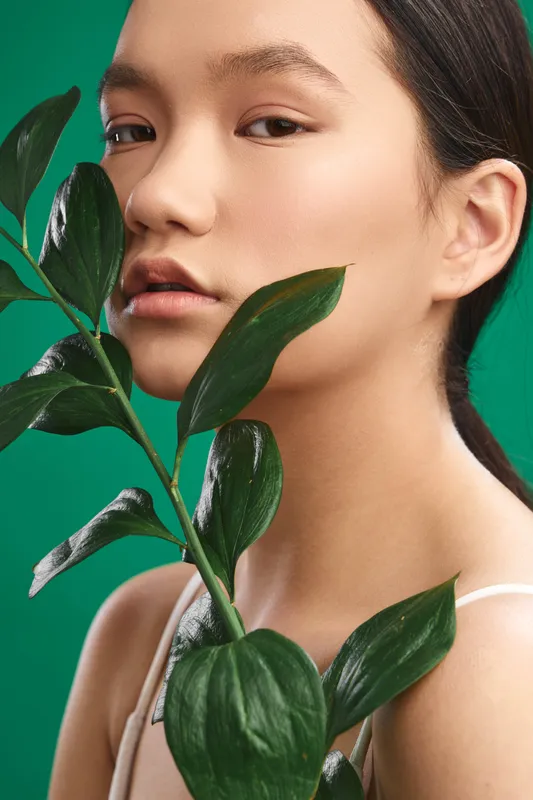 Asian woman gazes softly at the camera among lush leaves against a vibrant green backdrop, conveying calm beauty, modern style, and a natural, fresh aesthetic vibe.
