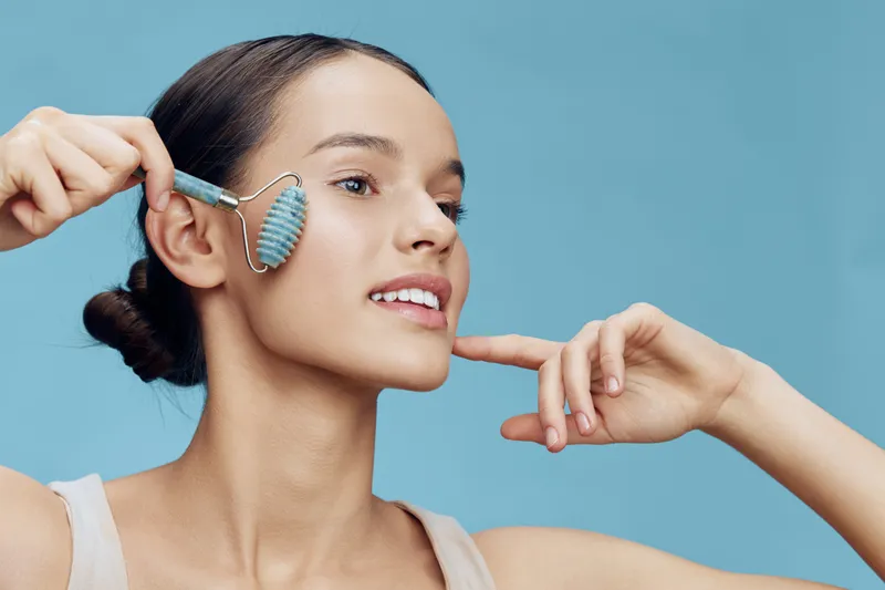 Young woman using a jade roller on her face, showcasing beauty and skincare routine with a serene expression against a pastel blue background