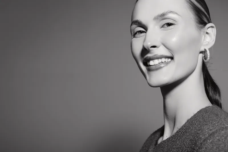 Smiling white woman with light skin and pulled back hair poses in studio with grey background. Concept for beauty, happiness, confidence, and natural portrait photography.