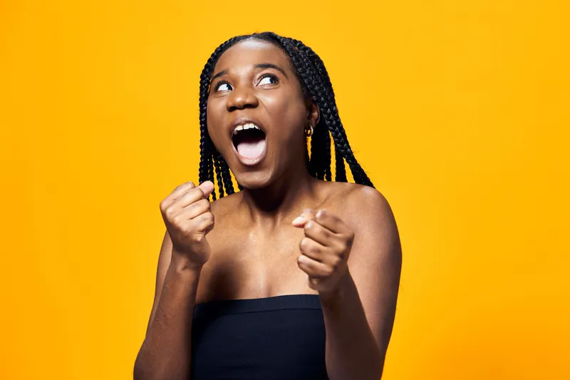 Excited young woman expressing joy with raised fists on a vibrant yellow background, showcasing enthusiasm and positivity in a modern style