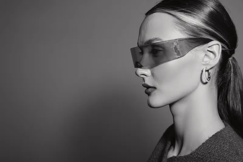 Profile of a light skinned Caucasian woman with long hair wearing futuristic glasses and earrings, monochrome photo with studio lighting and neutral background.