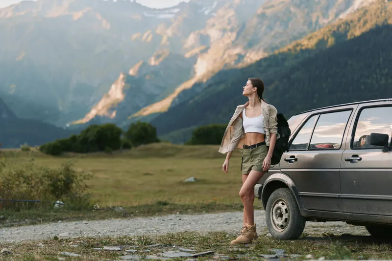 Woman stands beside a rugged car in a mountain meadow, wearing shorts and a cropped top with a jacket, embracing an adventurous outdoor lifestyle and wide horizon, wind in hair, sense of freedom,
