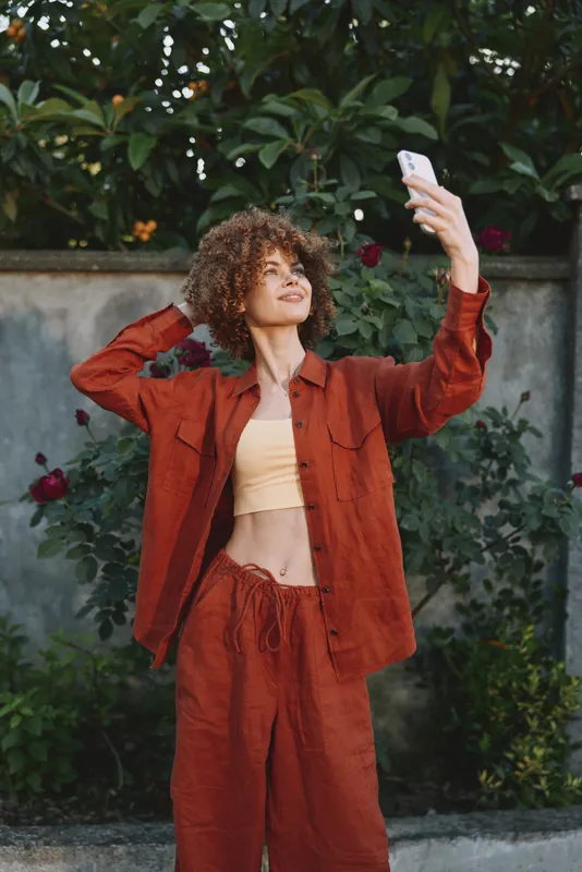 Cheerful woman outdoors, posing in a rust tracksuit, taking a selfie, warm sunlight, relaxed pose, vibrant greenery, casual lifestyle moment