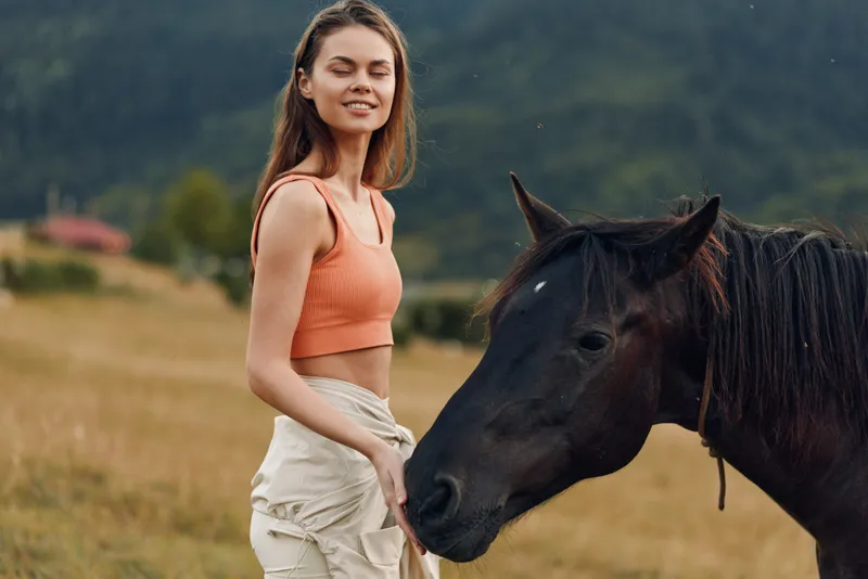 A relaxed woman stands beside a dark horse in an open countryside field. She wears a peach crop top and light shorts, smiling softly, enjoying calm outdoor companionship with the graceful animal.