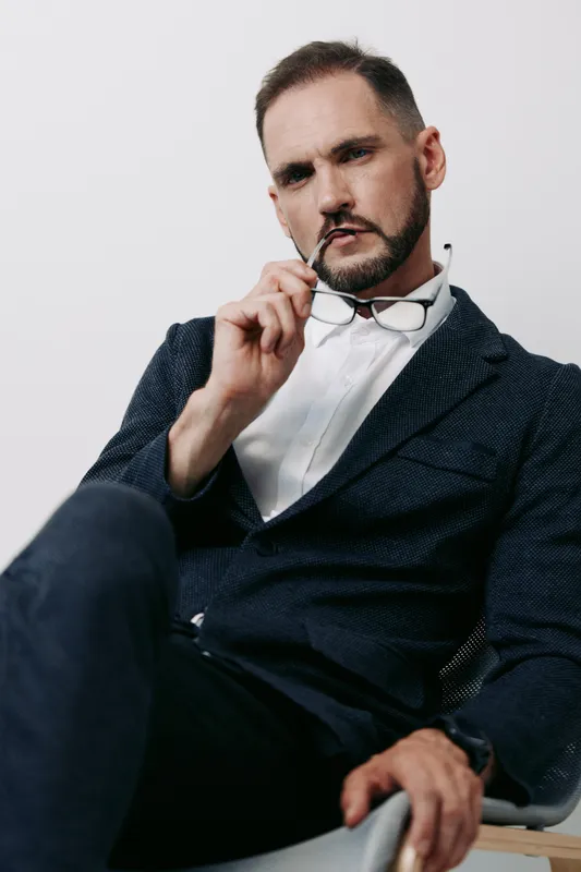 A professional man in a tailored suit sits with a confident gaze against a solid colored background, projecting focus, calm authority, and poised business demeanor.