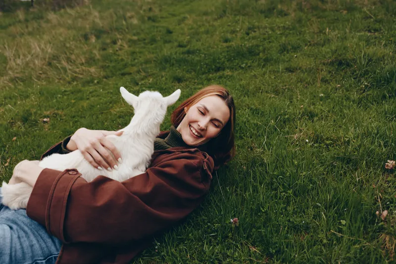 Smiling woman lying on grass, hugging a white goat kid, enjoying a gentle outdoor moment. Natural light creates a warm, relaxed mood, rural vibe, bonding, wellness, self care, cozy connection,