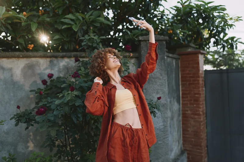 Young woman with curly hair in a red jacket raises her arm in a sunlit garden, smiling as roses frame a brick wall behind her, capturing a playful lifestyle moment