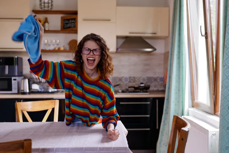 Cheerful woman with curly hair wearing a colorful striped sweater waves a blue glove over a kitchen table, bright window light, capturing a candid home cleaning moment.