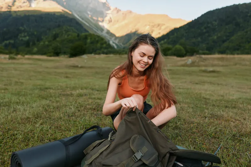 A cheerful woman outdoors in a grassy field packs a backpack beside a rolled mat with mountains in the background, hinting at a calm, adventurous outdoor experience.