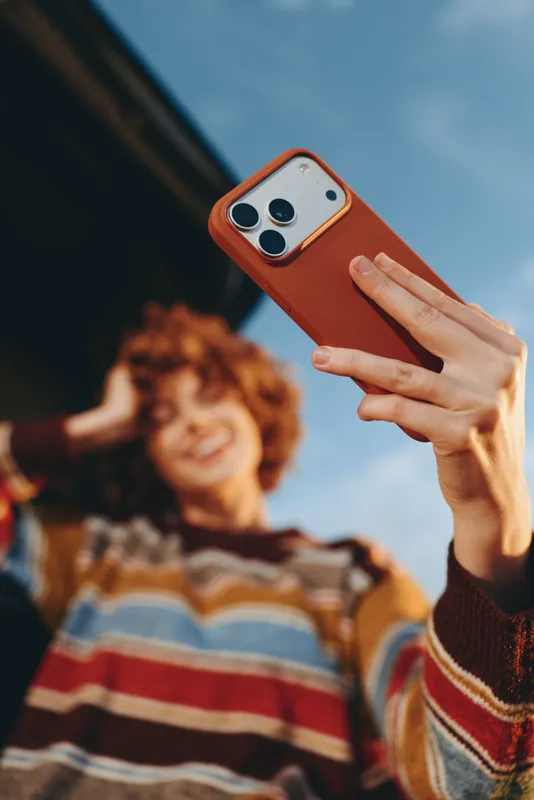 Woman lifestyle selfie outdoors, holding smartphone in hand, smiling in background, warm summer sky, casual striped sweater, soft natural light, relaxed candid moment, gentle breeze softly