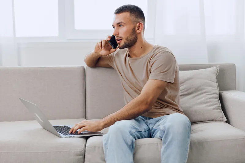 Engaged man working on a laptop while talking on a phone, enjoying the comfort of a cozy interior. Perfect for remote work and productivity themes.