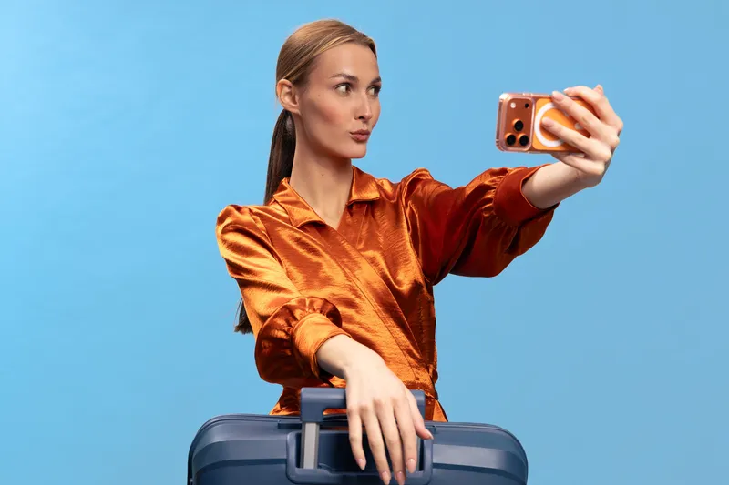 Young woman in orange satin blouse takes a selfie while holding a suitcase handle against blue background. Concept of travel, lifestyle, and modern communication with mobile phone.