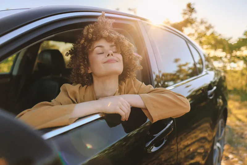 Young woman leans out of a car window, smiling with relaxed casual lifestyle, outdoor mood, portrait of happiness, sunshine and retro film tones, capturing a spontaneous moment of freedom