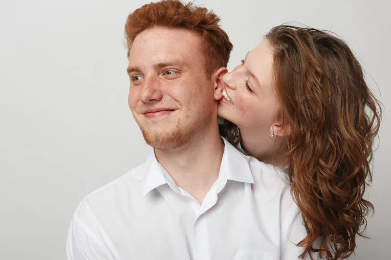 Happy young couple in white shirts share a joyful moment, woman smiling and leaning close to man against plain light background. Concept of love, friendship, and affection.