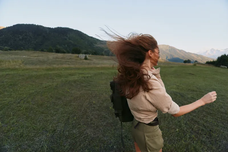 Woman with backpack in a grassy field, hair blown by wind, enjoying outdoor life. Lively mood of adventure, nature, and freedom under soft daylight.