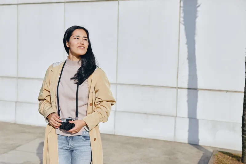 Woman with camera in casual fashion outdoors, standing confidently under bright sunlight, wearing beige coat and jeans, capturing candid street style and everyday moments.