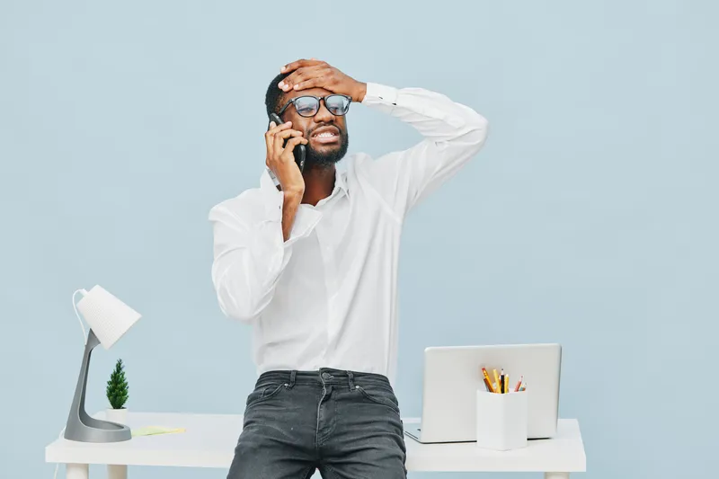 Frustrated young African American man with glasses, wearing white shirt, talking on smartphone while holding forehead, standing in office with modern desk, computer, and stationery, blue background. P
