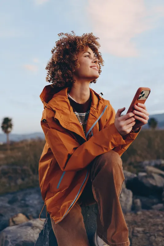 Woman lifestyle outdoors, orange jacket, curly hair, holding smartphone, smiling and looking at screen, warm sunset light, open landscape, natural colors, relaxed travel mood, fresh breeze