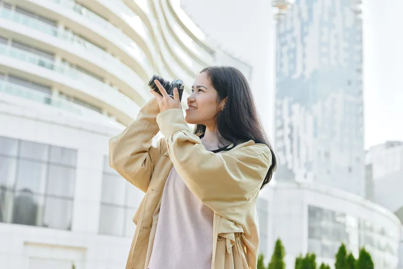 Camera in hand, smiling woman photographs urban landscape during daytime, wearing a beige coat in a modern city setting with tall buildings and clear sky.