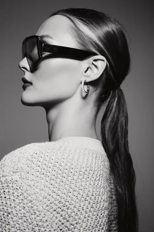 Stylish white woman with light skin wearing oversized sunglasses and hoop earrings in black and white studio portrait.