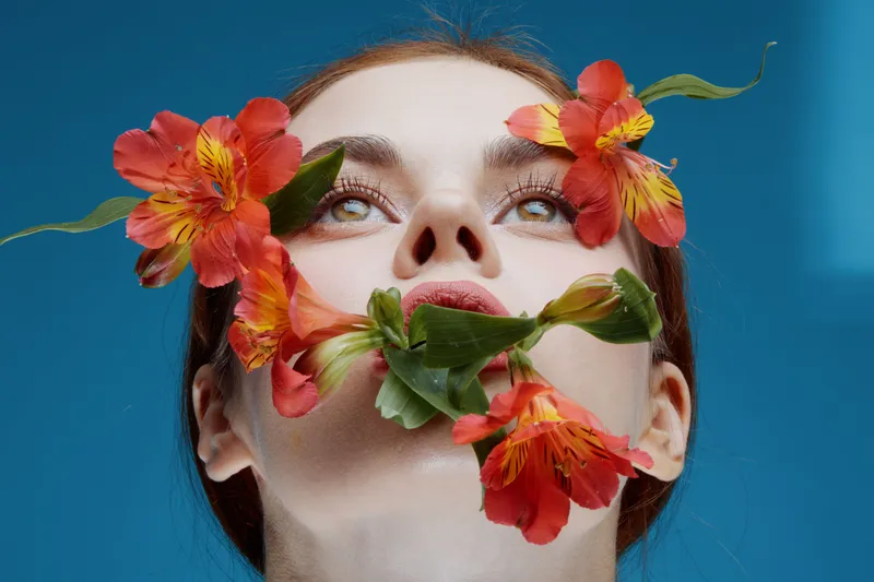 Young woman with flowers on her face, expressing tranquility and beauty against a blue background, representing nature and self care in a vibrant seasonal concept