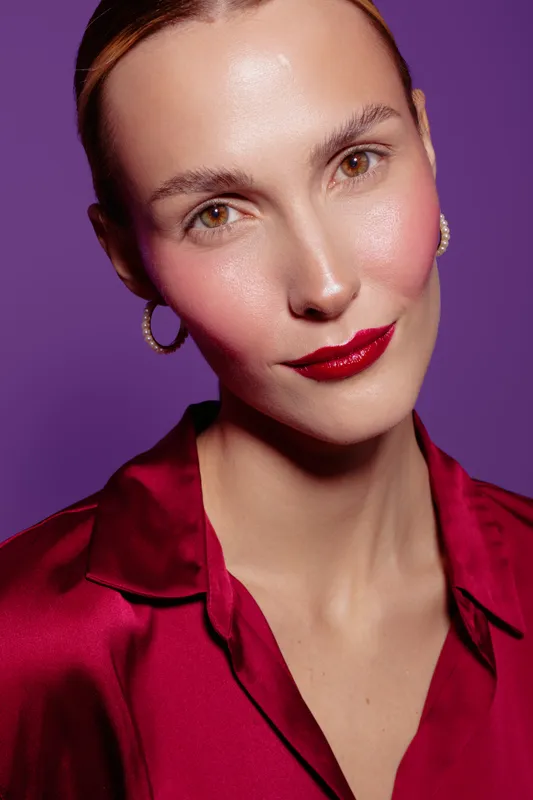 Close-up portrait of a confident woman with glossy red lips and smooth skin wearing a red satin blouse and hoop earrings against a purple background. Beauty and fashion concept.