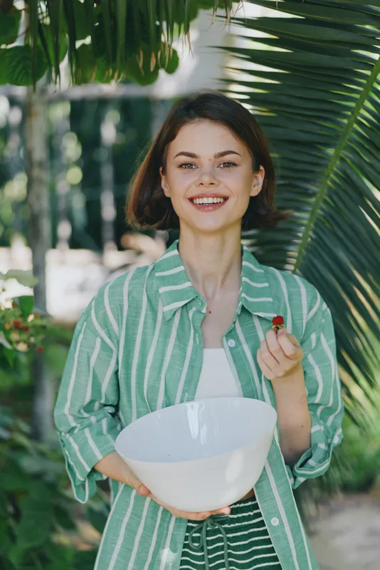 Smiling woman holding a white bowl in a lush tropical outdoor setting, wearing a green striped shirt.