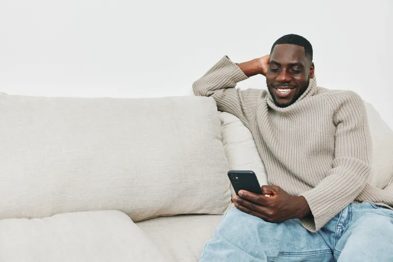 Relaxed Black man smiling while using a smartphone on a cozy couch, showcasing casual style and comfort in a modern living space