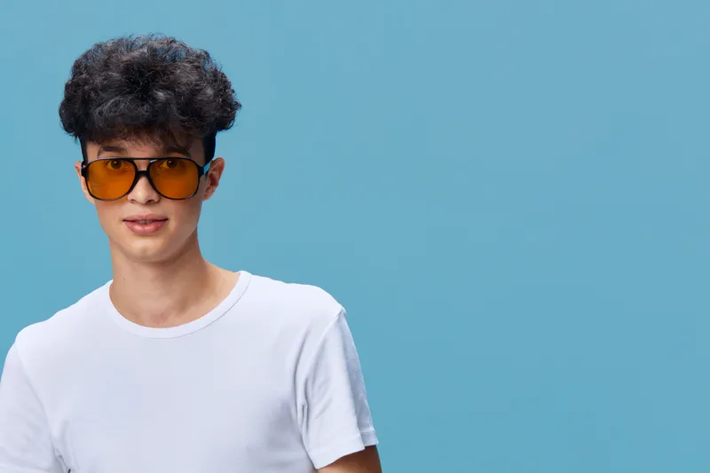 young man wearing oversized sunglasses and white t-shirt against blue background smiling confidently, casual style, trendy look, summer fashion vibe