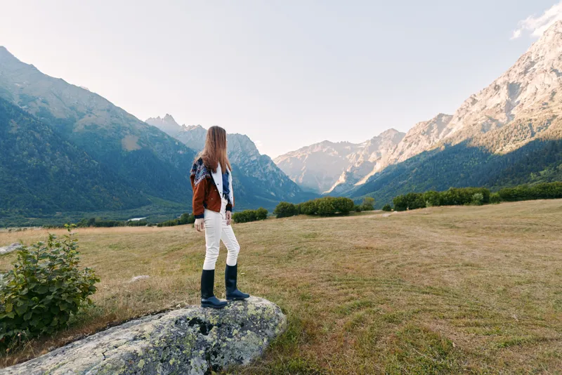 Woman standing on rock in mountain valley hiking with boots and jacket, looking toward distant mountains. Landscape and nature scene for outdoor adventure, solo travel and exploration.