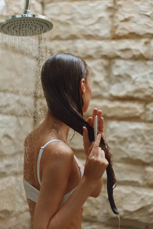 Woman washing hair under outdoor shower with wet skin and long dark hair, natural light, rustic stone wall background, relaxed moment in warm weather.