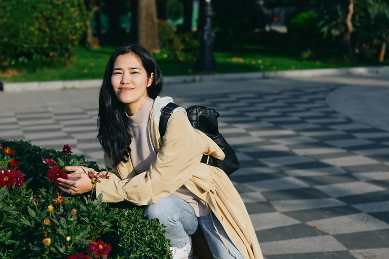 Woman kneeling beside vibrant flowers in an outdoor park wearing a beige coat and backpack, soft smile, morning light, candid lifestyle shot for nature and travel use