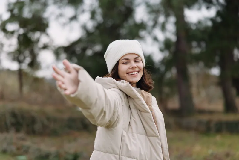 Woman outdoors in a light winter jacket and beanie, arms outstretched with a bright smile in a park setting, evoking joy, warmth and freedom.