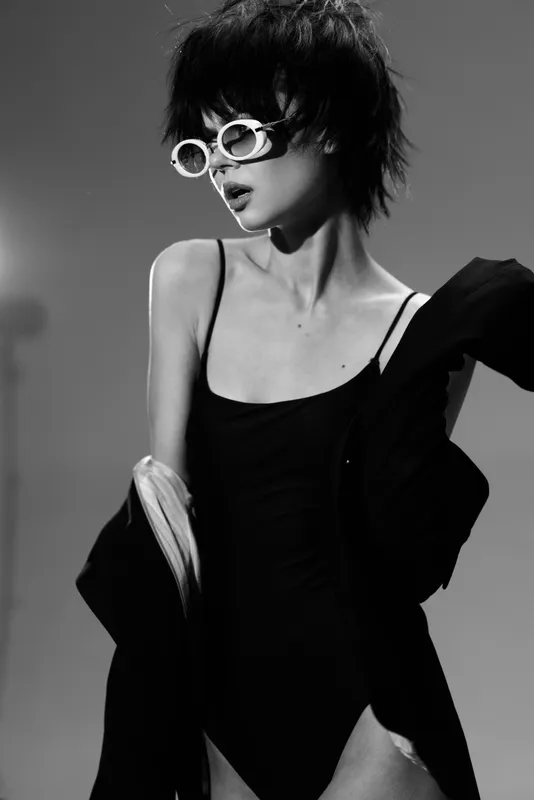Stylish woman wearing sunglasses and black outfit poses in artistic black and white fashion portrait with dramatic lighting.
