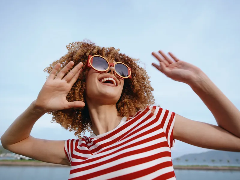 Joyful young woman with curly hair wearing a red and white striped shirt and round sunglasses, raising her hands in a carefree pose against a bright blue sky.