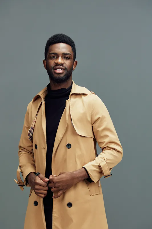 Stylish African male model in a beige trench coat, smiling against a muted green background, showcasing modern fashion trends with confidence