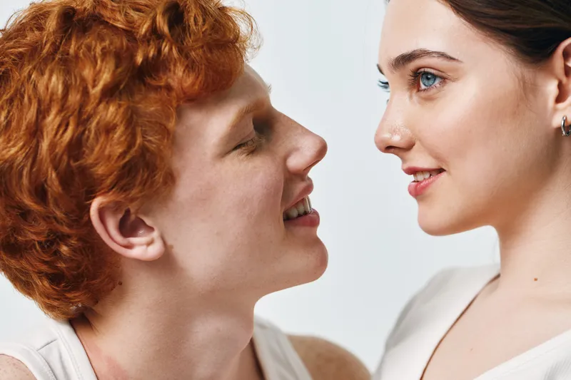 Portrait of young couple with joyful expressions looking at each other closeup. Smiling man with red hair and woman with blue eyes in intimate moment on light background.