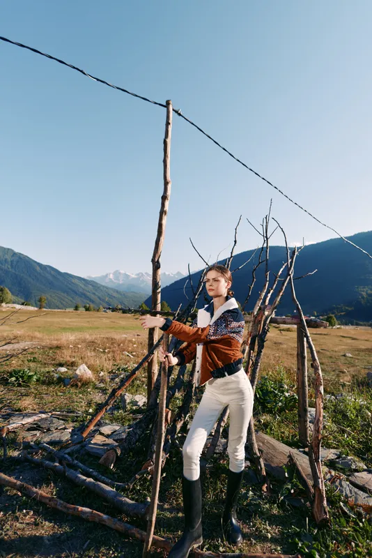 woman fence mountains field boots jacket rural outdoors portrait of young model leaning on handmade wooden fence in alpine meadow, casual riding style, natural light and distant peaks