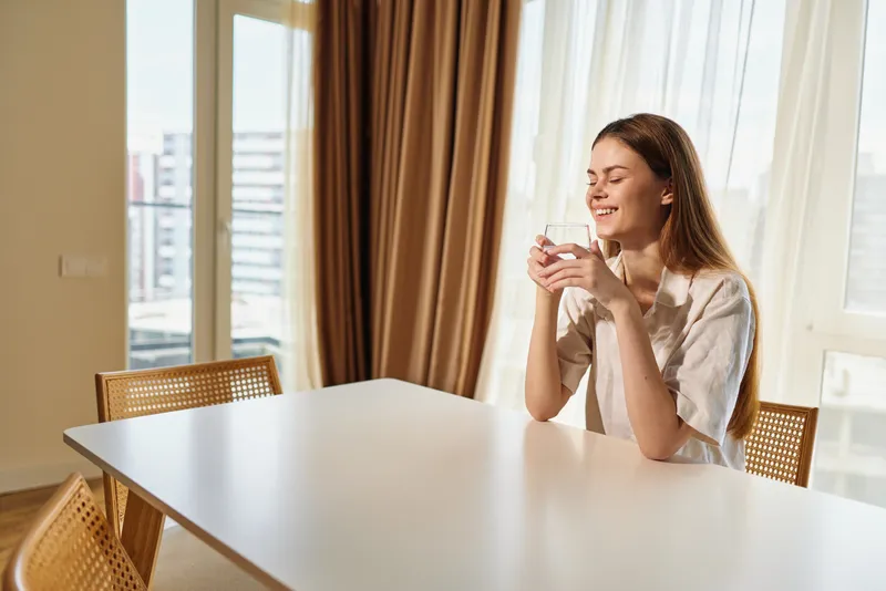 Woman sitting at table in bright morning sunlight, relaxed at home holding a cup and smiling, casual breakfast moment in cozy kitchen interior with warm natural light.