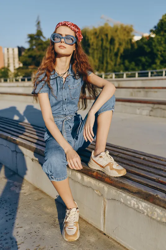 Lifestyle woman in modern boho-western denim street style wearing oversized sunglasses and sneakers, outdoor urban setting with film color effect, casual and trendy look.