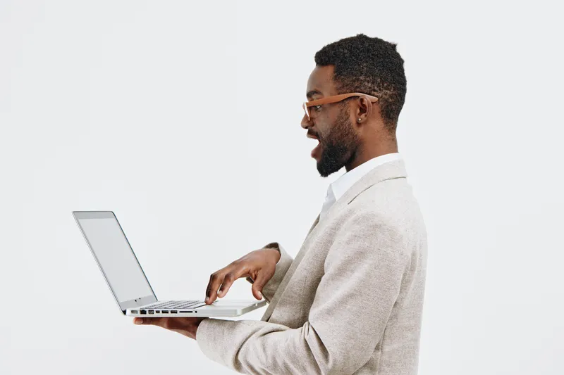 Stylish young African-American man wearing glasses and beige blazer working on a laptop, standing against a plain white background. Creative professional, technology, business, casual, modern look, in