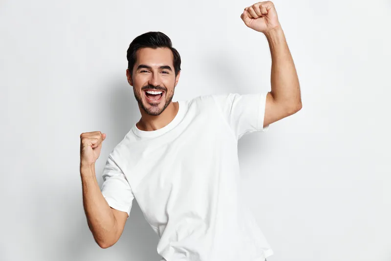 Happy smiling man celebrating success with arms raised and a joyful expression, wearing a casual white t-shirt, isolated on a simple light background.