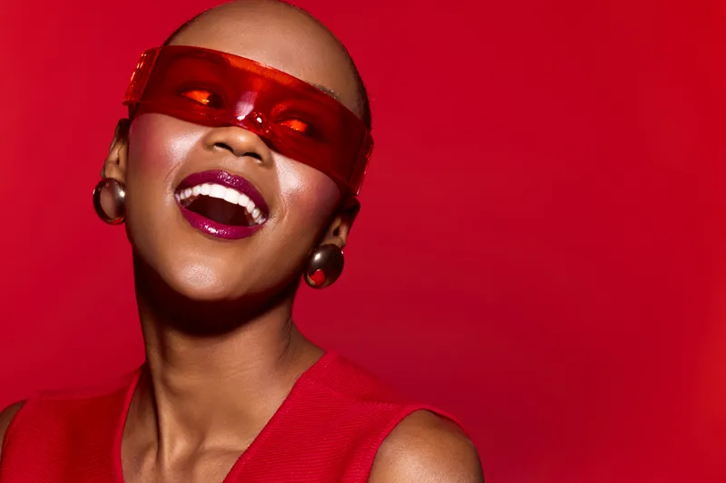 Happy African American woman with dark skin wearing red sunglasses and dress smiles brightly against a red background. Concept of fashion, joy, and vibrant lifestyle in studio lighting.